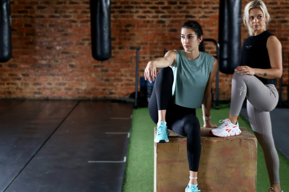 Two women in a gym setting, one sitting on a box and the other standing, with boxing bags in the background.
